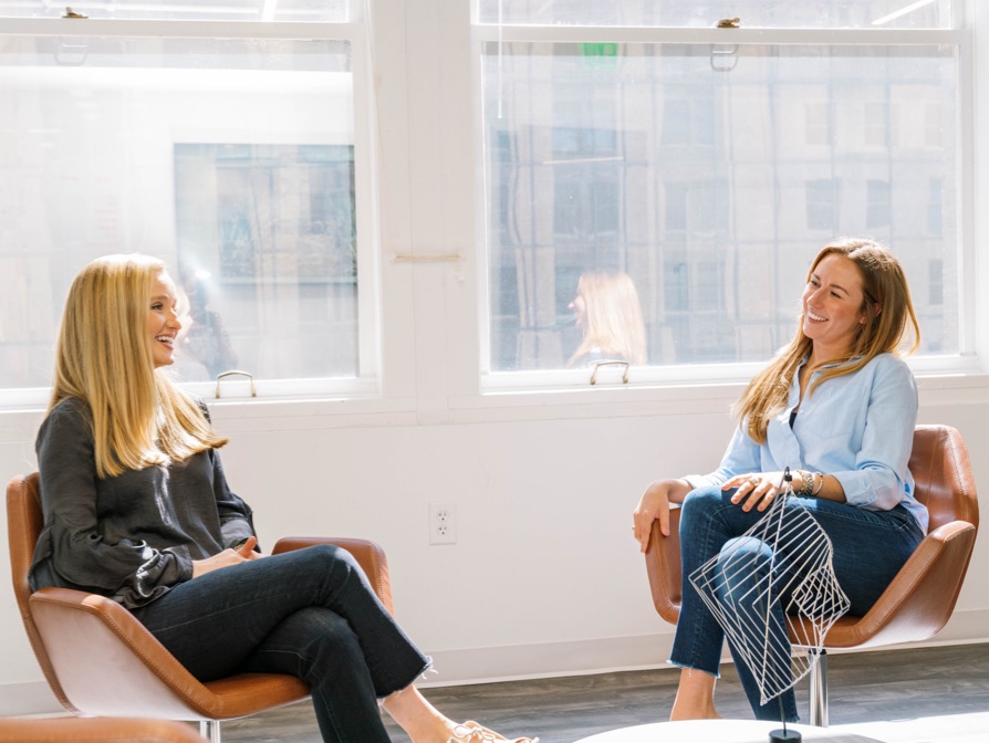 a photo of 2 women siting in chairs next to a sunny window smiling at each-other